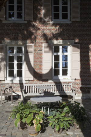 Close up of Atelier Pierre Culot frontage, a bench, an iron table surrounded with plants, the shadow of a tree.