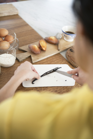 A person cutting a vanilla pod on a chopping board next to madeleines and a cup of coffee.