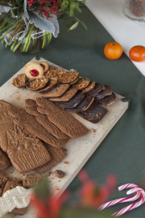 Verschillende maten Sinterklaas en enkele Dandoy koekjes op een snijplank, met daarnaast clementines, bloemen en suikerriet.